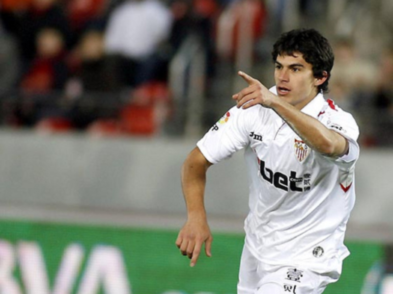 Un jugador de fútbol en acción, con una camiseta blanca y roja, y una bandera en el brazo. El fondo muestra una grada con espectadores y un anuncio de 'BVA' en el suelo.