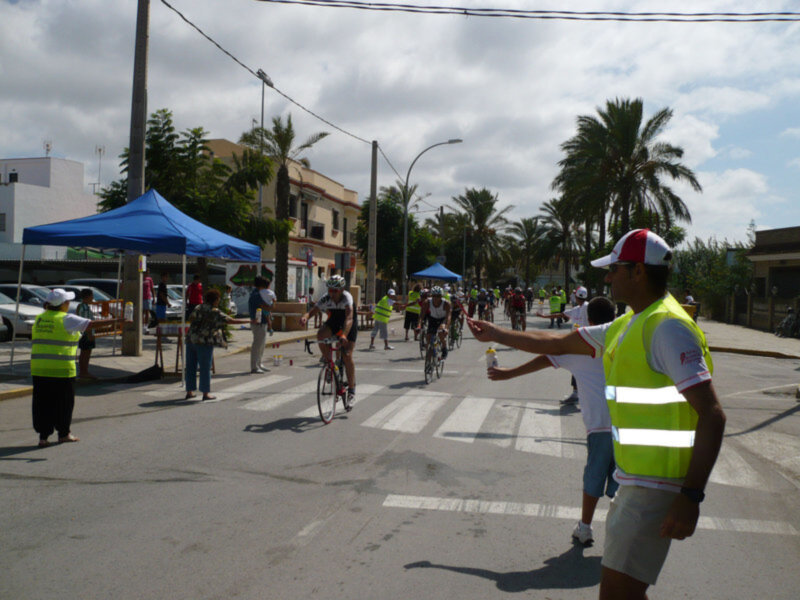 Ciclistas participando en una carrera urbana bajo un cielo parcialmente nublado, con voluntarios y espectadores en el borde de la carretera.