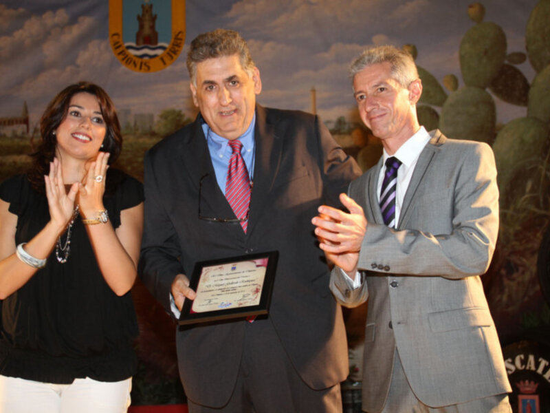 Dos personas celebran la entrega de un reconocimiento, con una mujer sonriendo y un hombre sosteniendo un diploma, frente a un fondo con una pintura de cactus y un edificio.