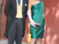 Una pareja elegante posa frente a un edificio con una pared roja. La mujer lleva un vestido verde con un hombro descubierto y guantes blancos, mientras que el hombre está vestido con un traje formal y corbata azul.