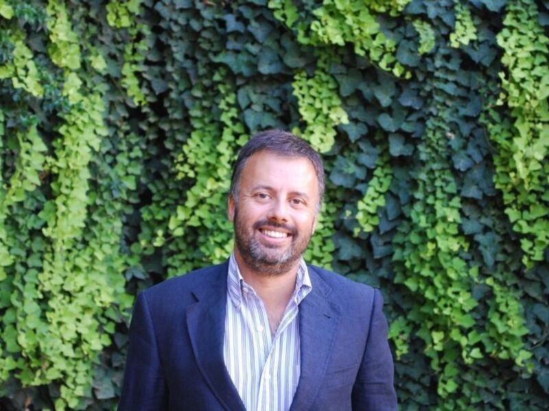 Un hombre con barba y bigote sonríe frente a una pared de plantas verdes.