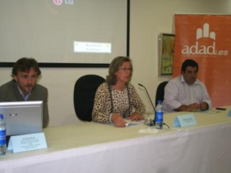 Una conferencia con tres personas sentadas en una mesa, con una pantalla de presentación en el fondo y un cartel con la marca 'adad.es' en la pared.