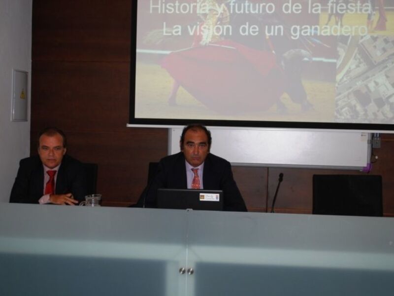 Dos hombres sentados en una mesa con laptops, frente a un proyector que muestra una imagen de toros y texto sobre la historia y futuro de la fiesta, con el título "La visión de un ganadero".