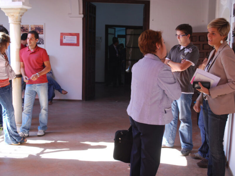 Una escena en un edificio con varias personas intercambiando información, uno de ellos sostiene un bolso y una persona con una chaqueta beige sostiene un folleto.