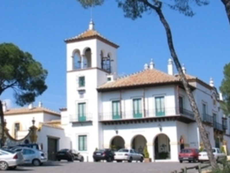 Vista frontal de un edificio colonial con torre y balcones, rodeado de árboles y coches en el estacionamiento.