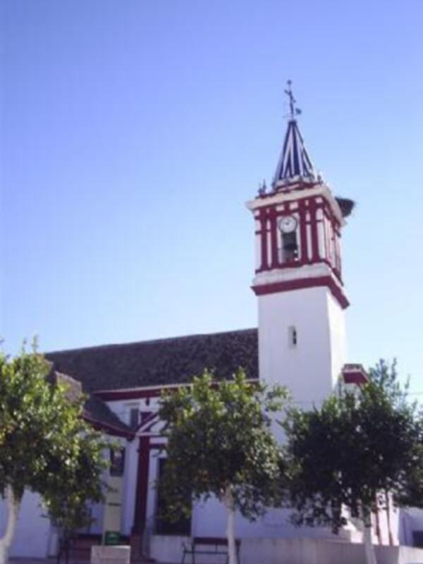 Iglesia con torre blanca y roja en un pueblo español, rodeada de árboles y bancos.