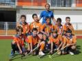 Un grupo de niños vestidos con uniformes naranjas y azules posando para una foto en un campo de hockey. Algunos niños sostienen palos de hockey, y el entrenador está sentado entre ellos. La imagen muestra un ambiente deportivo y de equipo.
