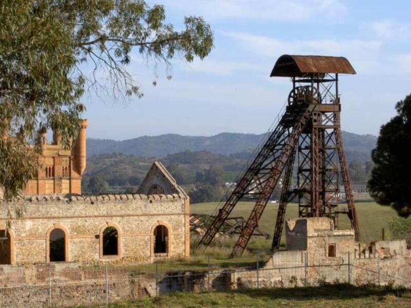 Torre de control de una mina abandonada, con una estructura de hierro oxidada y una edificación de ladrillo rojo en el fondo, rodeada de vegetación y un paisaje rural.