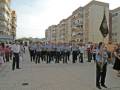 Fotografía de un grupo de uniformados en una plaza pública, con edificios residenciales al fondo. Un oficial sostiene un estandarte con insignias.