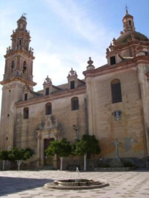 Iglesia histórica con torre y cúpulas en un patio de plaza con fuente y árboles.