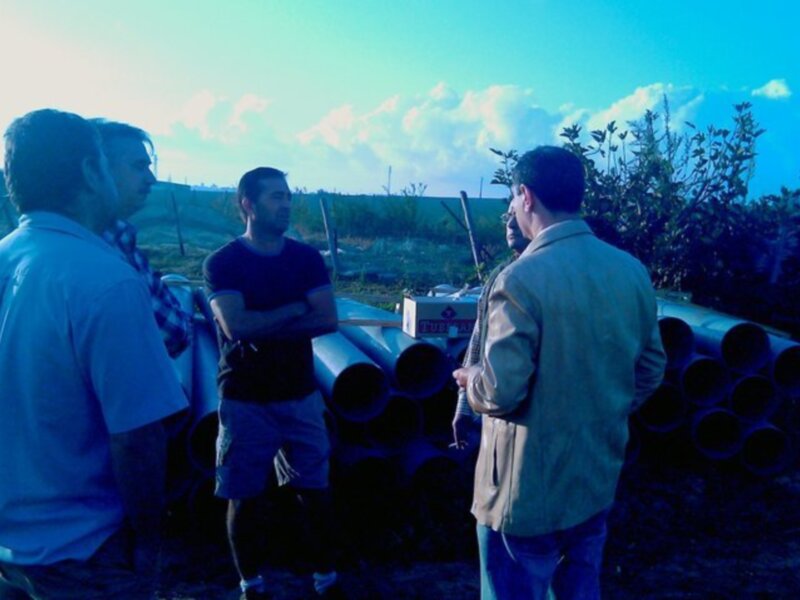 Tres hombres conversan en una zona de construcción con tubos y herramientas al fondo.