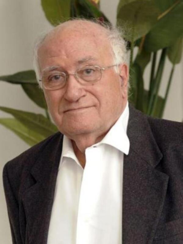 Retrato de un hombre con cabello gris, gafas y traje oscuro, posando frente a una planta verde.