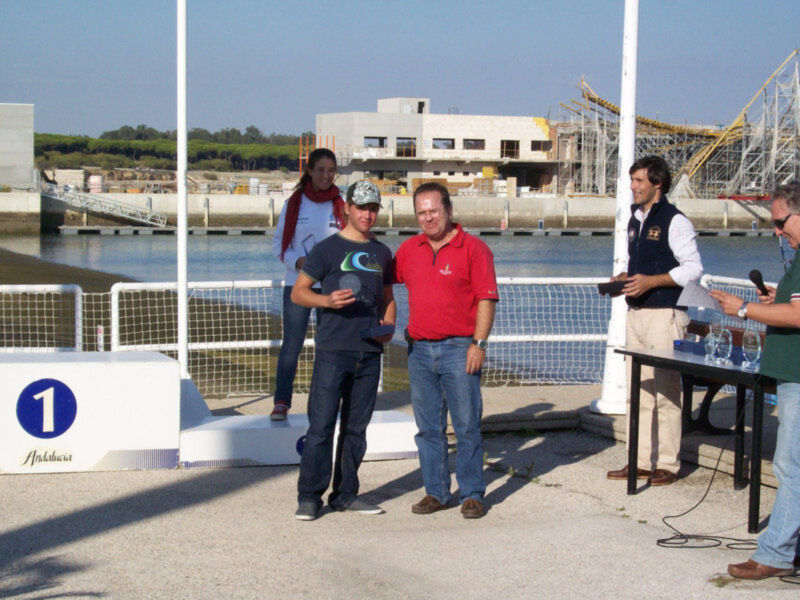 Premiación en un evento deportivo junto al agua, con un fondo de construcción y un podio con el número 1.