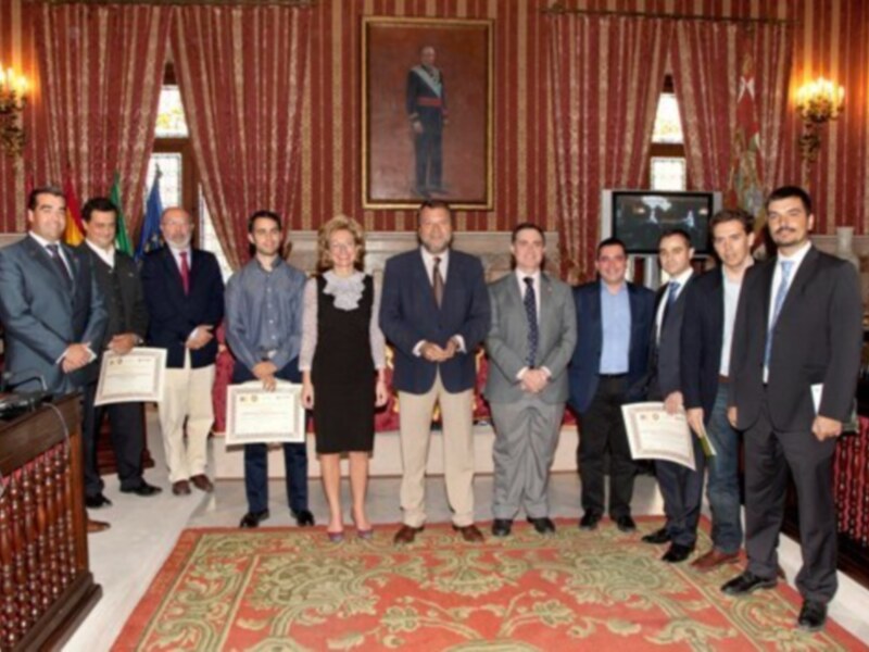 Gente en un salón con cortinas rojas y una foto de un hombre en el fondo, posando con diplomas. El ambiente parece formal y ceremonial.