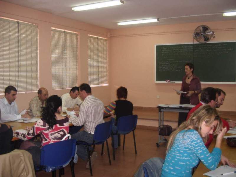 Clase de adultos en un aula con profesora frente a la pizarra y estudiantes trabajando en sus escritorios.