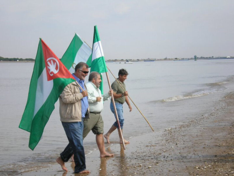 Tres personas caminan por la orilla del mar sosteniendo banderas de diferentes colores y diseños.