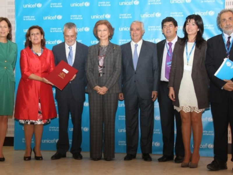 Una ceremonia de entrega de un reconocimiento en el contexto de UNICEF, con varios miembros de la organización y una mujer en el centro, sosteniendo un documento rojo.