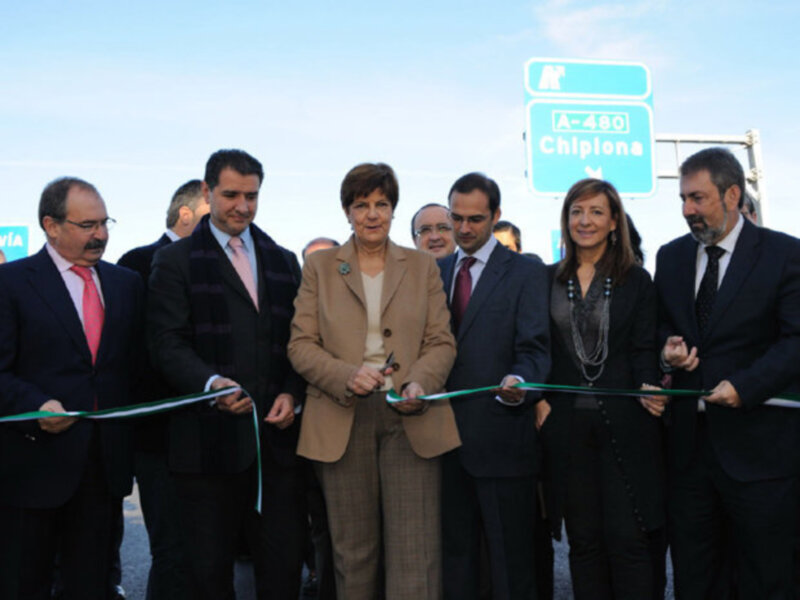 Una celebración de inauguración con una mujer en el centro cortando un lazo de cinta, rodeada de personas en trajes formales. En el fondo, se puede ver un letrero que indica 'A-480 Chilpanca'.