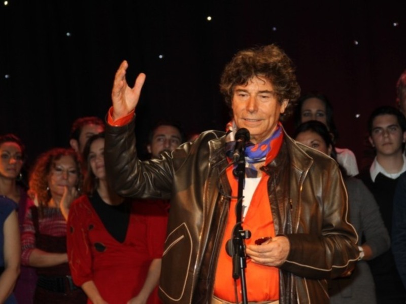Un hombre con una chaqueta de cuero y una camisa roja con un lazo de papel en la boca, saludando a una audiencia en un escenario con estrellas en el fondo.