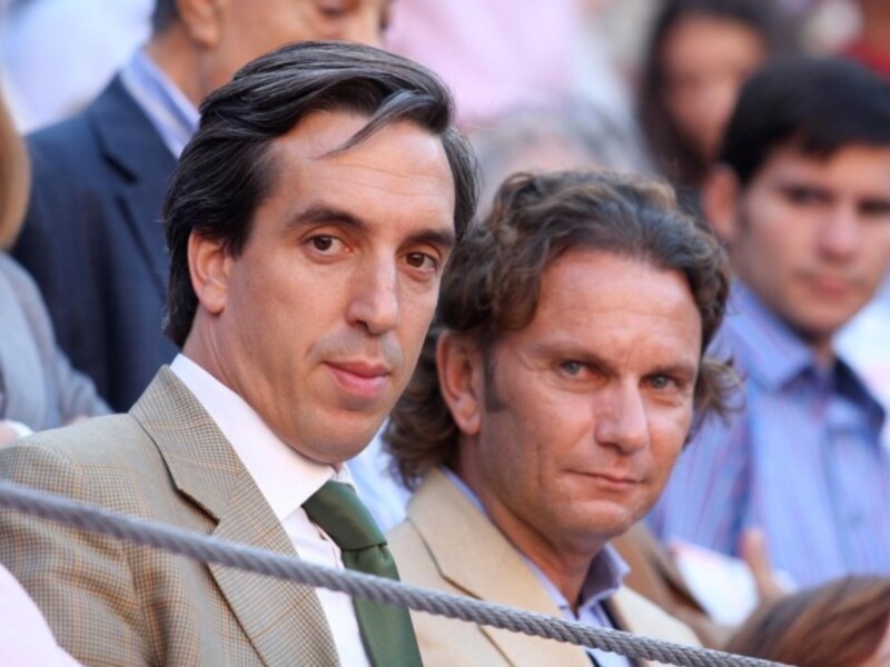Dos hombres en una reunión formal, uno con una camisa blanca y corbata verde, el otro con una camisa azul claro y corbata blanca, ambos con una expresión seria.