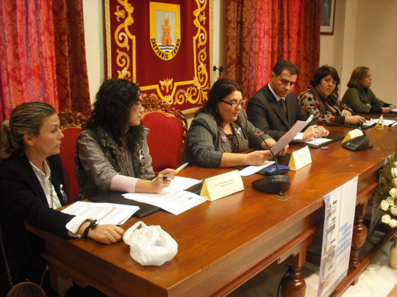 Reunión de trabajo en una sala con cortinas rojas y escudo de armas en el fondo, participantes sentados en una mesa con documentos y micrófonos.