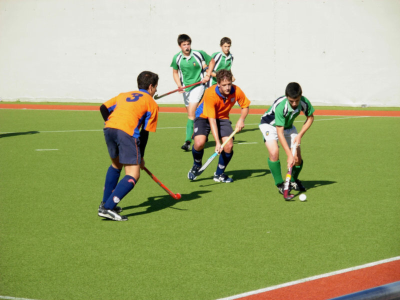 Jóvenes jugando hockey sobre hierba en un campo verde con una pared blanca en el fondo.