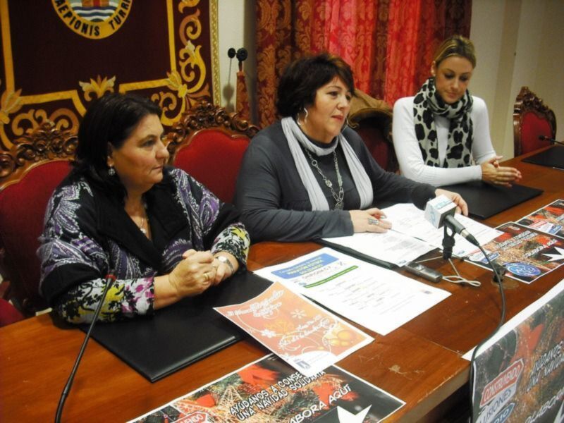 Tres mujeres participan en una conferencia de prensa en un salón con decoración formal y carteles de eventos navideños en la mesa.