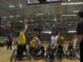 Un jugador en silla de ruedas lanza un balón hacia el aro durante un partido de baloncesto en una cancha deportiva. Otros jugadores y espectadores observan la acción desde los asientos del estadio.