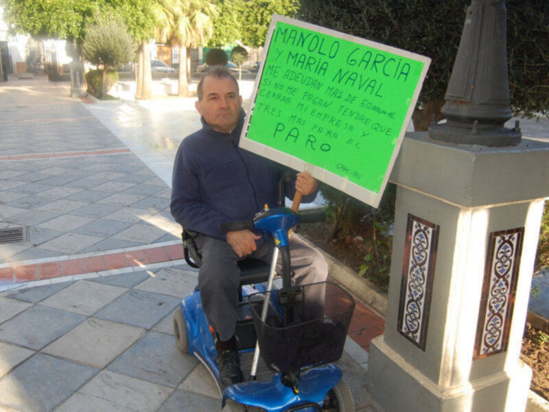 Un hombre en silla de ruedas sosteniendo una pancarta en la calle. La pancarta dice: 'Manolo García y María Naval. Me adeudan más de 8.000 euros. Si no me pagan más de 8.000 euros, iré a paro.'