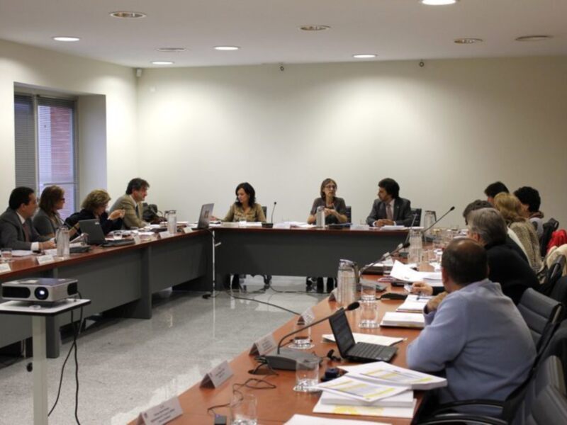 Sala de reuniones con personas sentadas en mesas, laptops y documentos en la mesa central, iluminada con luces de techo.