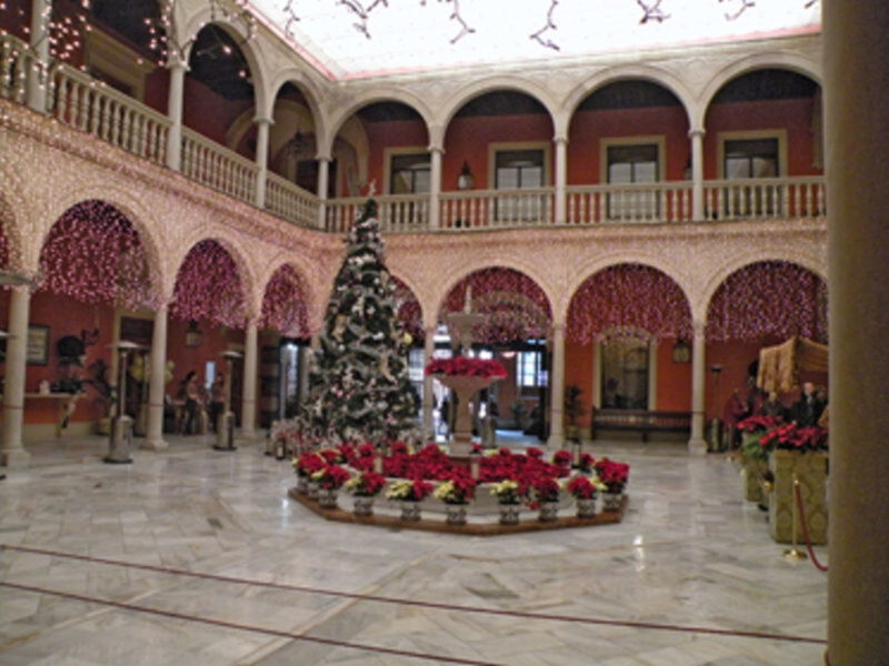 Interior de un patio decorado con árbol de Navidad y flores, con arcos y balcones en un ambiente festivo.