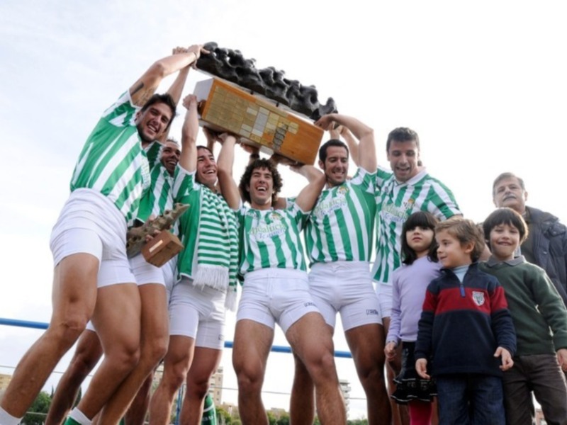 Un equipo de fútbol celebra una victoria, sosteniendo un trofeo en alto, mientras sus jugadores y aficionados celebran juntos en un campo de juego.