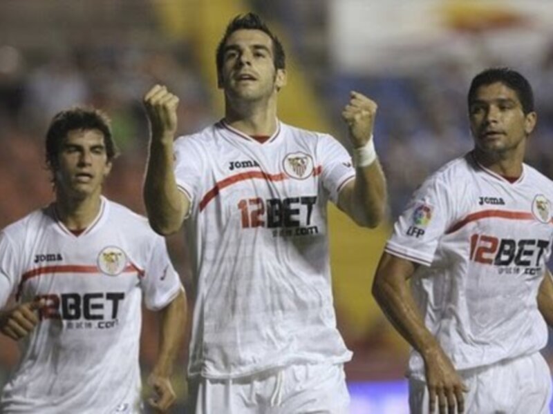 Tres jugadores de fútbol celebrando en el campo de juego, con camisetas blancas y rojas de la marca Joma.