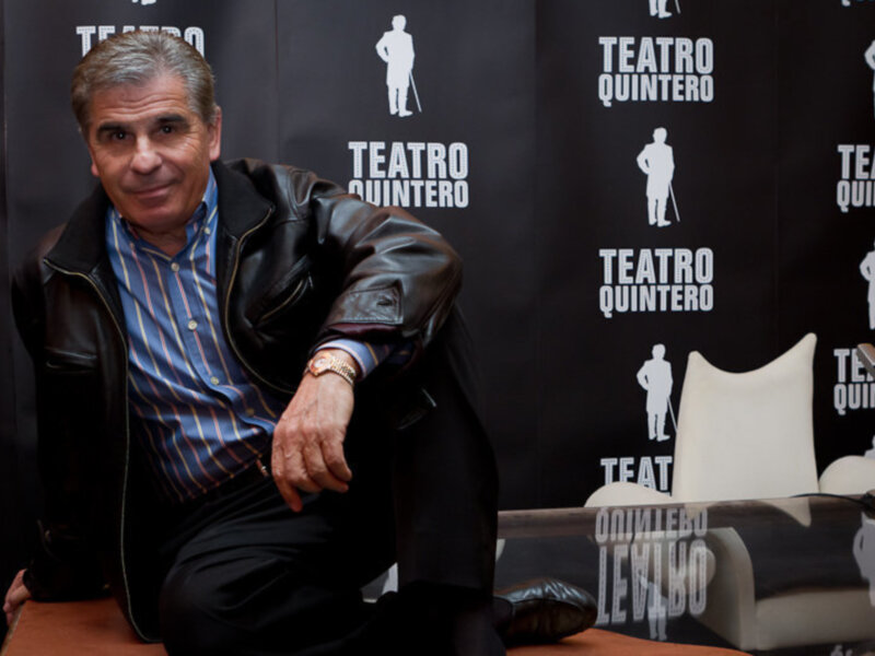 Un hombre sentado en una silla blanca frente a un fondo con el logo del Teatro Quintero, posando con una sonrisa.