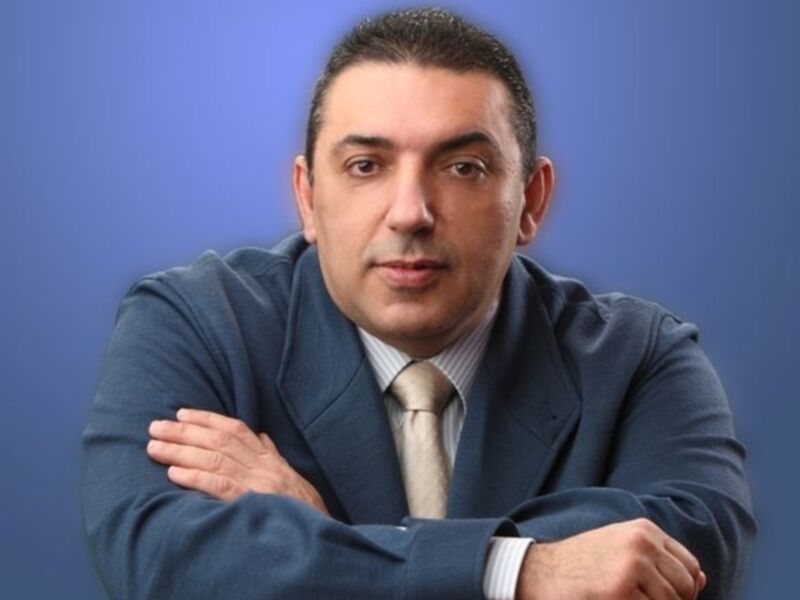 Hombre con traje y corbata, sentado con los brazos cruzados, fondo azul claro.