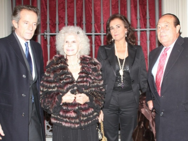Una imagen de tres personas vestidas formalmente, posiblemente en una celebración o evento importante, con fondos de cortinas rojas y una puerta de hierro en el fondo.