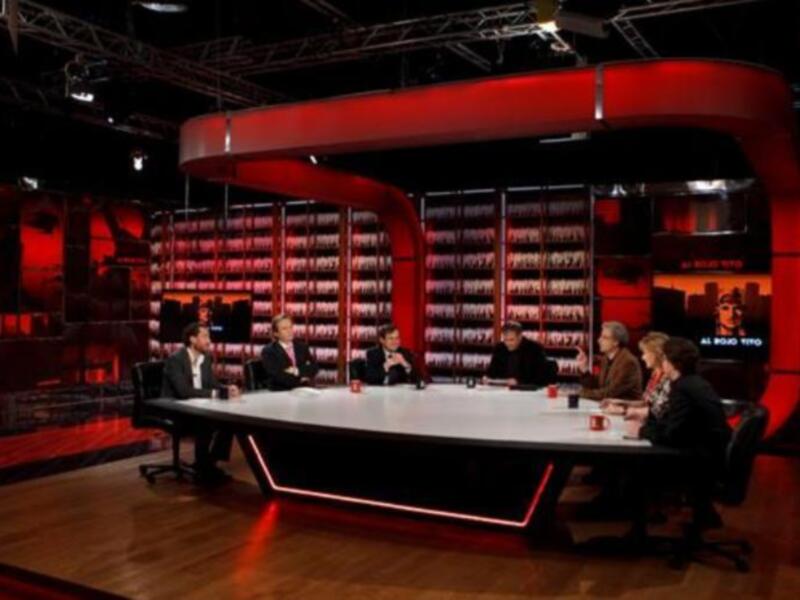 Una sala de conferencias con una mesa grande y sillas alrededor. Los asistentes están sentados y discutiendo. La sala tiene una iluminación roja en el fondo.