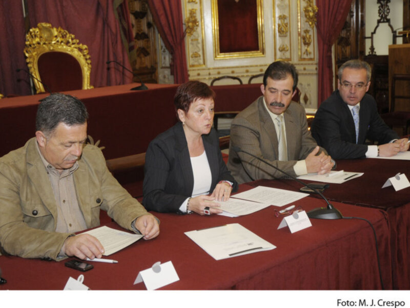 Tres personas sentadas en una mesa, con documentos y teléfonos en frente de ellos, en un ambiente formal con cortinas rojas y decoración dorada. Foto: M. J. Crespo.