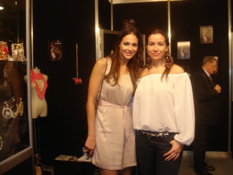 Dos mujeres posan juntas en un evento de moda, con vestimenta elegante y joyas de fondo.