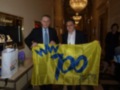 Dos hombres sostienen una bandera amarilla con el texto "100 años Cádiz CF 2010-2010". La imagen se encuentra en un interior elegante con una lámpara de cristal y un espejo.