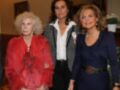 Tres mujeres posan juntas en un ambiente formal. La mujer de la izquierda lleva una chaqueta roja, la central tiene un traje blanco y negro con una camisa blanca, y la de la derecha usa un vestido azul con un cinturón dorado. La escena parece ser una reunión o evento formal.