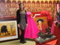 Una mujer posa junto a una escultura de un toro y un cuadro con un personaje en traje de charro, ambos en una exposición artística.