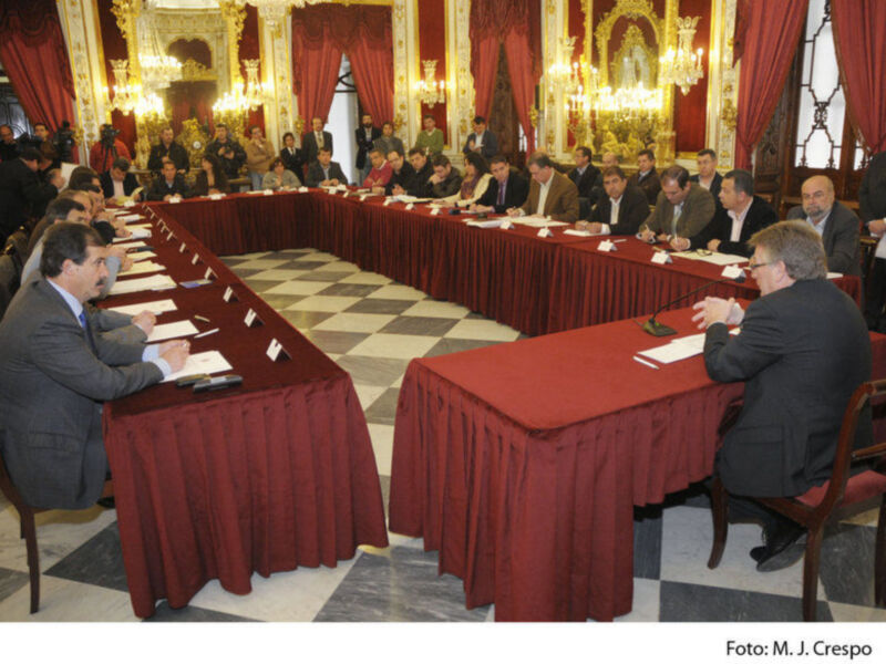 Reunión formal en un salón decorado con cortinas rojas y lámparas, con participantes sentados en largas mesas de rojo.