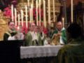En una celebración religiosa, sacerdotes vestidos en hábitos verdes se encuentran frente a un altar con velas encendidas y flores en el fondo. La atmósfera es solemnemente reverente, indicando un acto de culto o ceremonia.