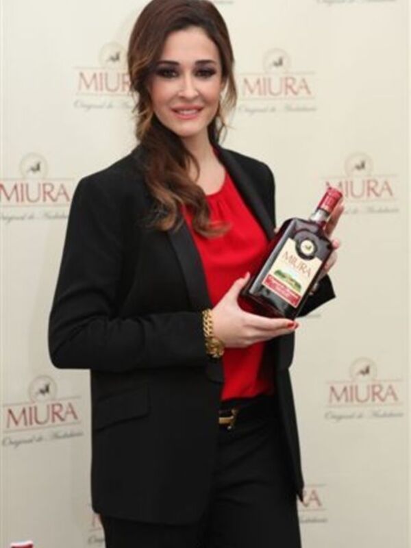 Una mujer en traje negro y camisa roja sostiene una botella de licor Miura en una presentación de producto.