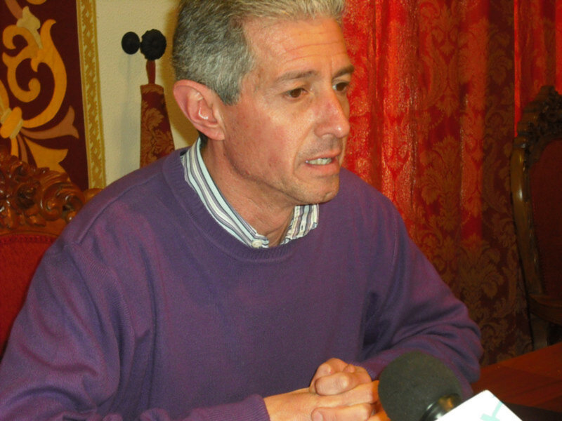 Persona hablando con micrófono, vestida con suéter morado en un entorno interior con cortinas rojas y detalles ornamentales.