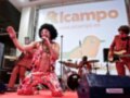 Artista en escenario con banda, vestido colorido y afro, actuando en evento Alcampo.