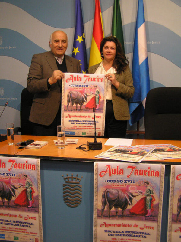 Un hombre y una mujer sostienen un cartel de la Escuela Municipal de Tauromaquia de Jerez, con banderas española y europea al fondo.