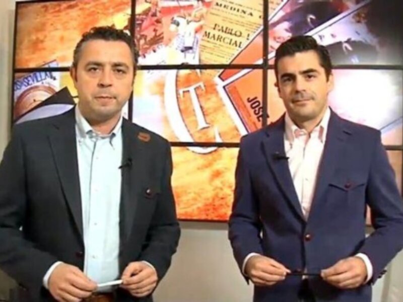 Presentadores en un estudio de televisión con fondo de noticias y publicidad.