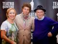En el Teatro Quintero, tres personas sonrientes posan frente a un fondo con la palabra "Teatro Quintero". La persona de la izquierda lleva una camiseta con un diseño de árbol, el centro está vestido con un traje beige y una camisa con estampado, mientras que la persona de la derecha usa un suéter azul y una gorra.